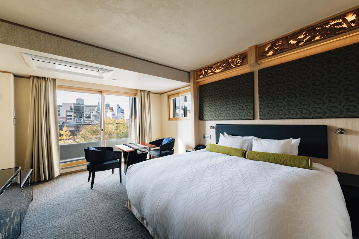 A hotel room with a large bed, a desk, a chair, and a view of the city through large windows.