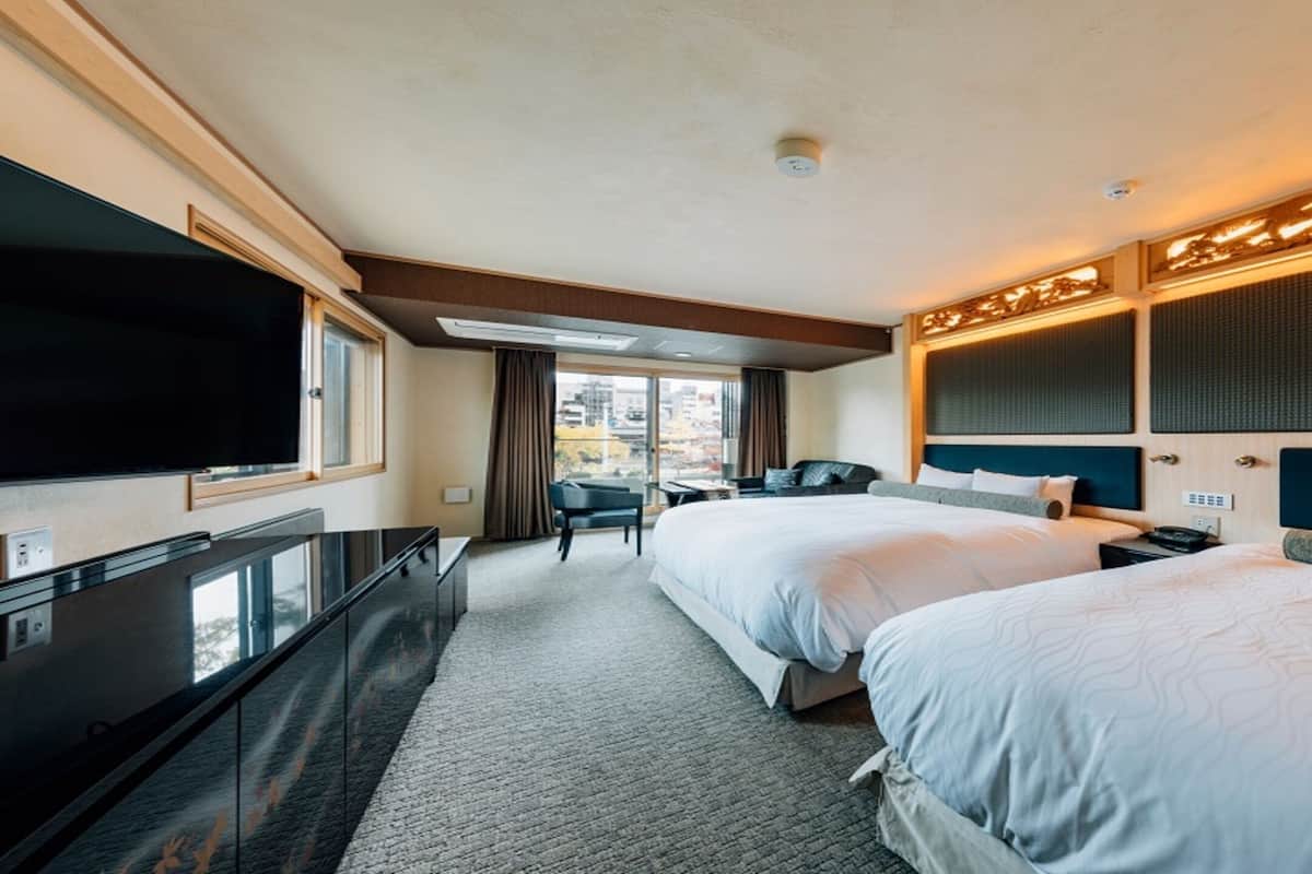 A hotel room with two beds, a TV, a desk, and a view of the city.