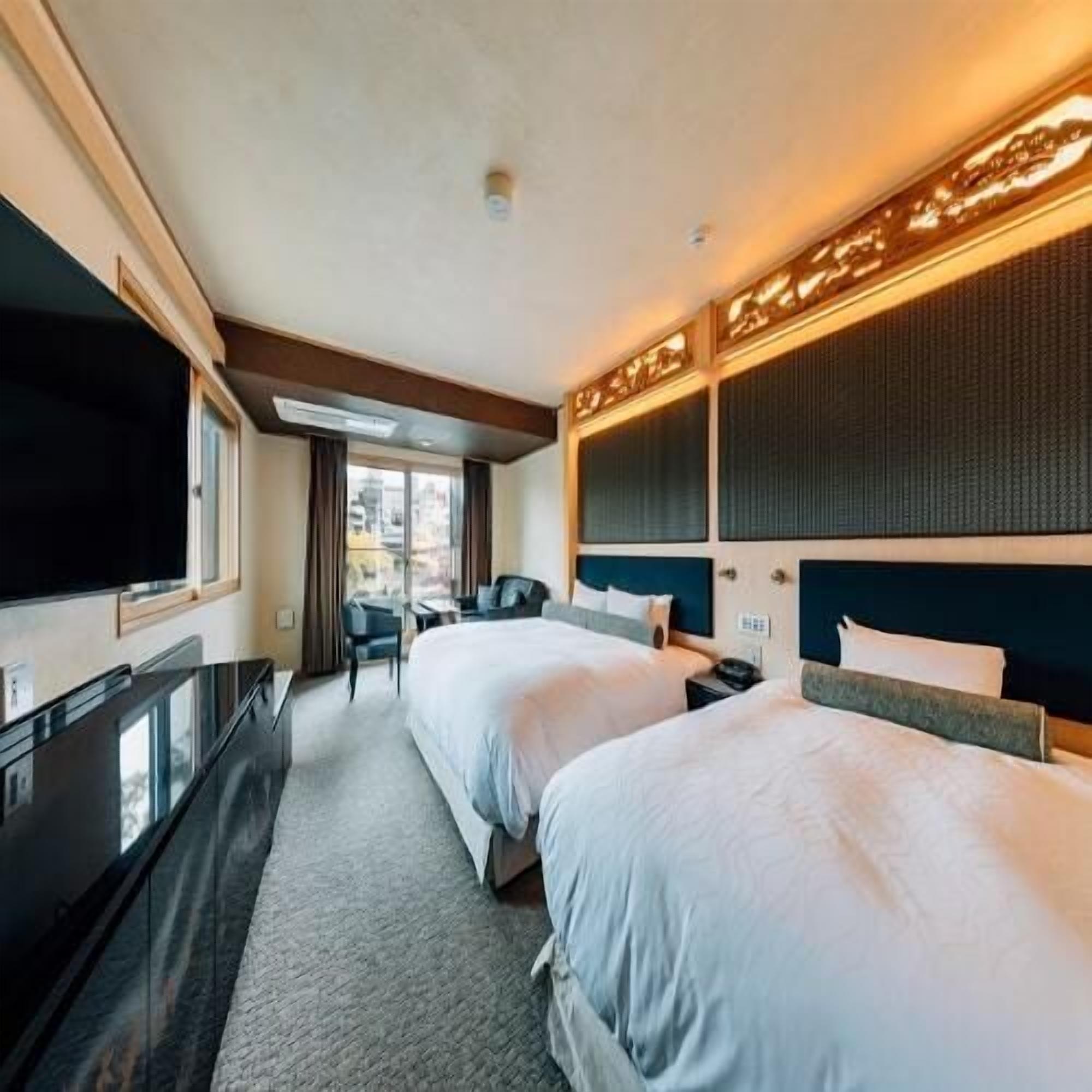 A hotel room with two beds, a desk, a TV, and a large window.