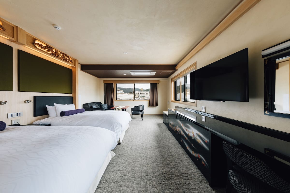 A hotel room with two beds, a desk, a TV, and a window with a city view.