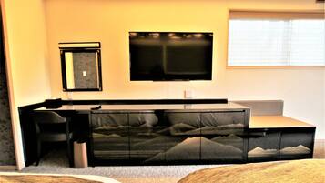 A hotel room with a sofa bed, a desk, a chair, a television, and a window with blinds.