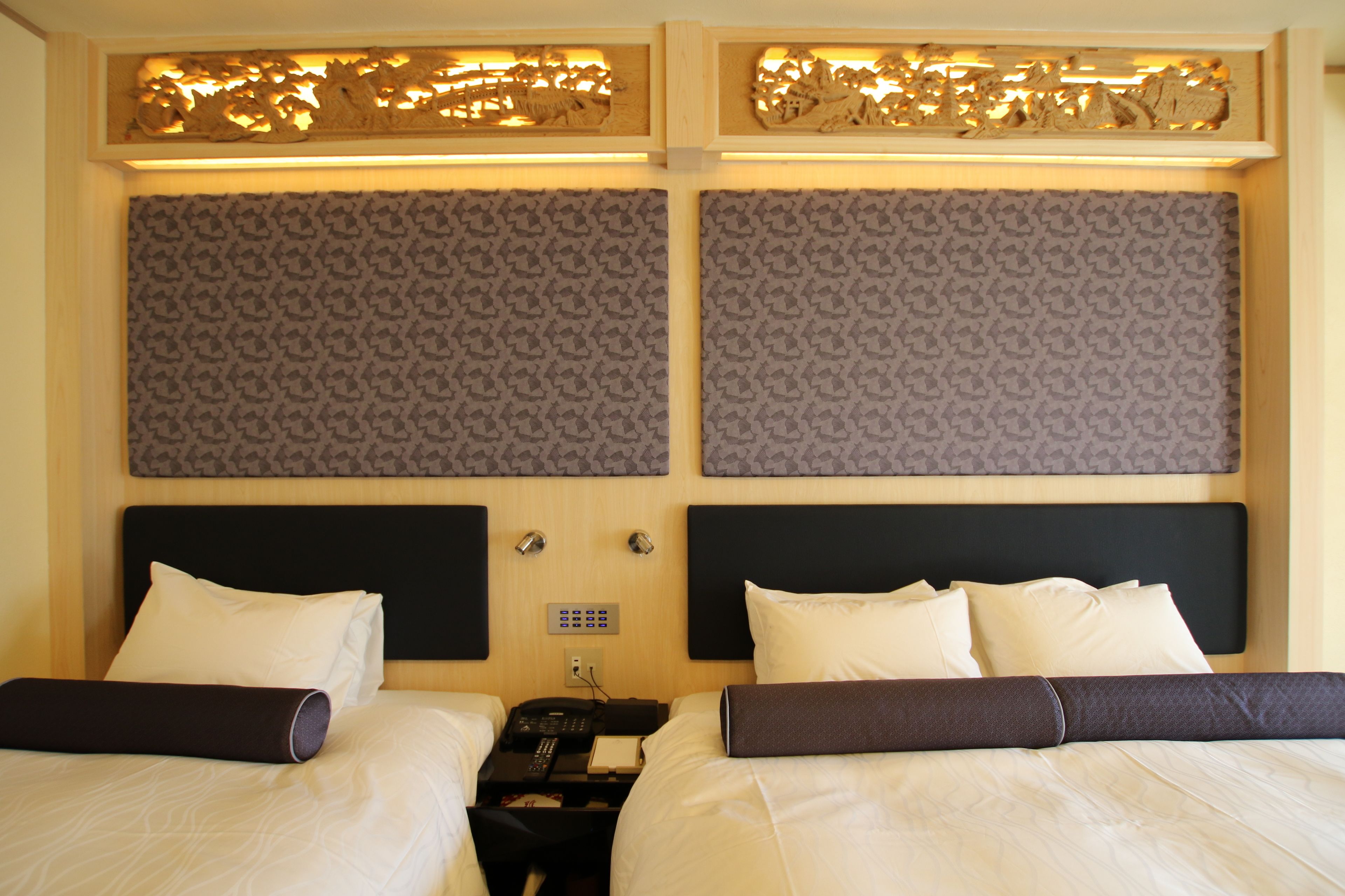 Habitación de hotel con dos camas, cabecero de madera, pared con diseño y una mesita de noche pequeña con un teléfono y un libro.