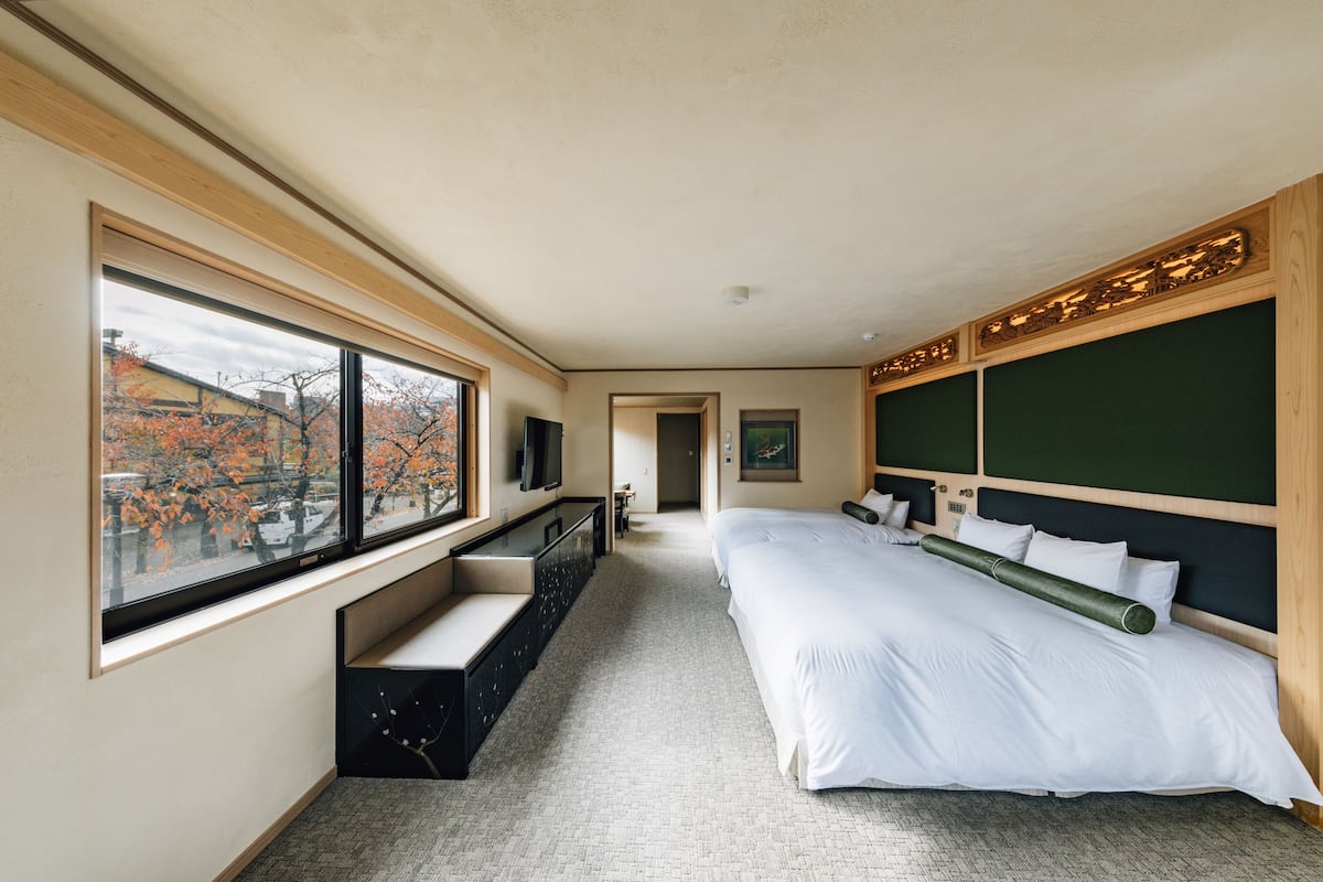 A modern hotel room with a large bed, a built-in bench, a TV, and a view of autumn trees through large windows.