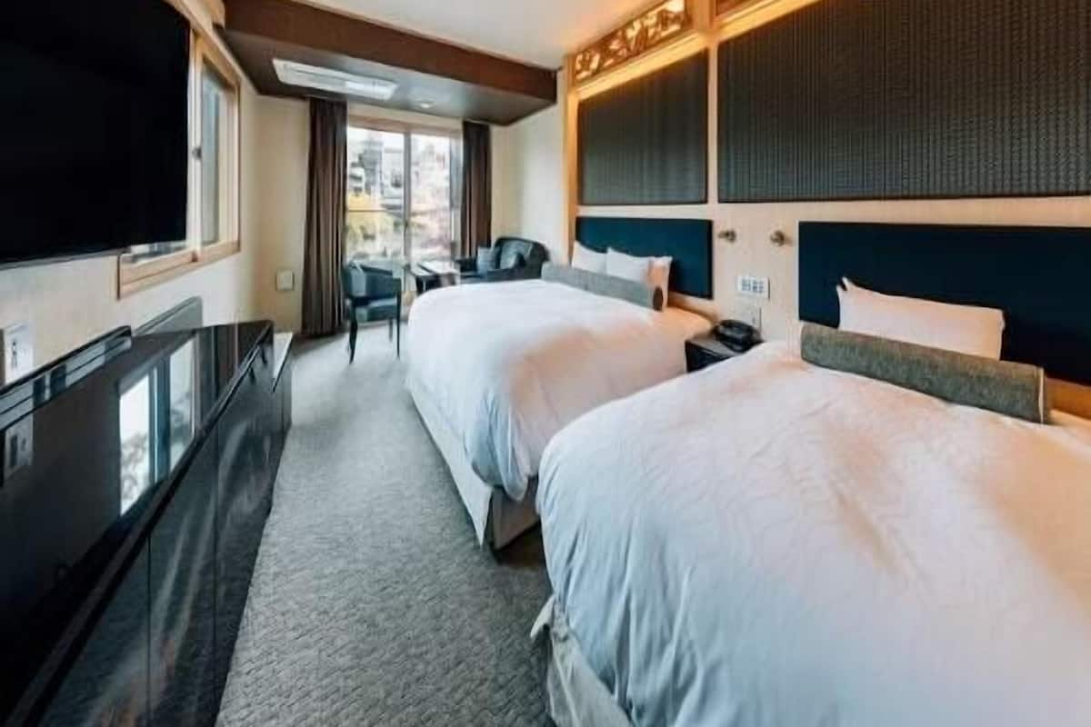 A hotel room with two beds, a desk, a flat-screen TV, and a large window.