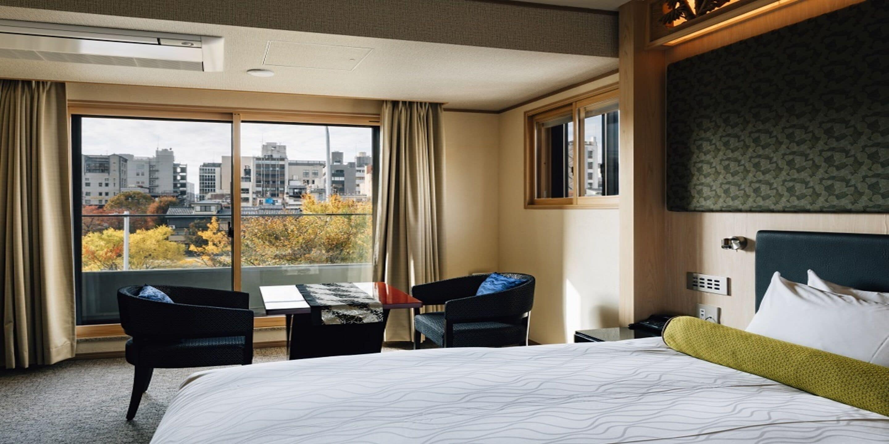 Une chambre d’hôtel avec un grand lit, un bureau avec deux chaises, une vue sur la ville grâce à de grandes fenêtres et un panorama urbain visible à l’extérieur.
