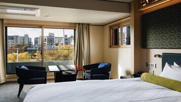 Une chambre d’hôtel avec un grand lit, un bureau avec deux chaises, une vue sur la ville grâce à de grandes fenêtres et un panorama urbain visible à l’extérieur.