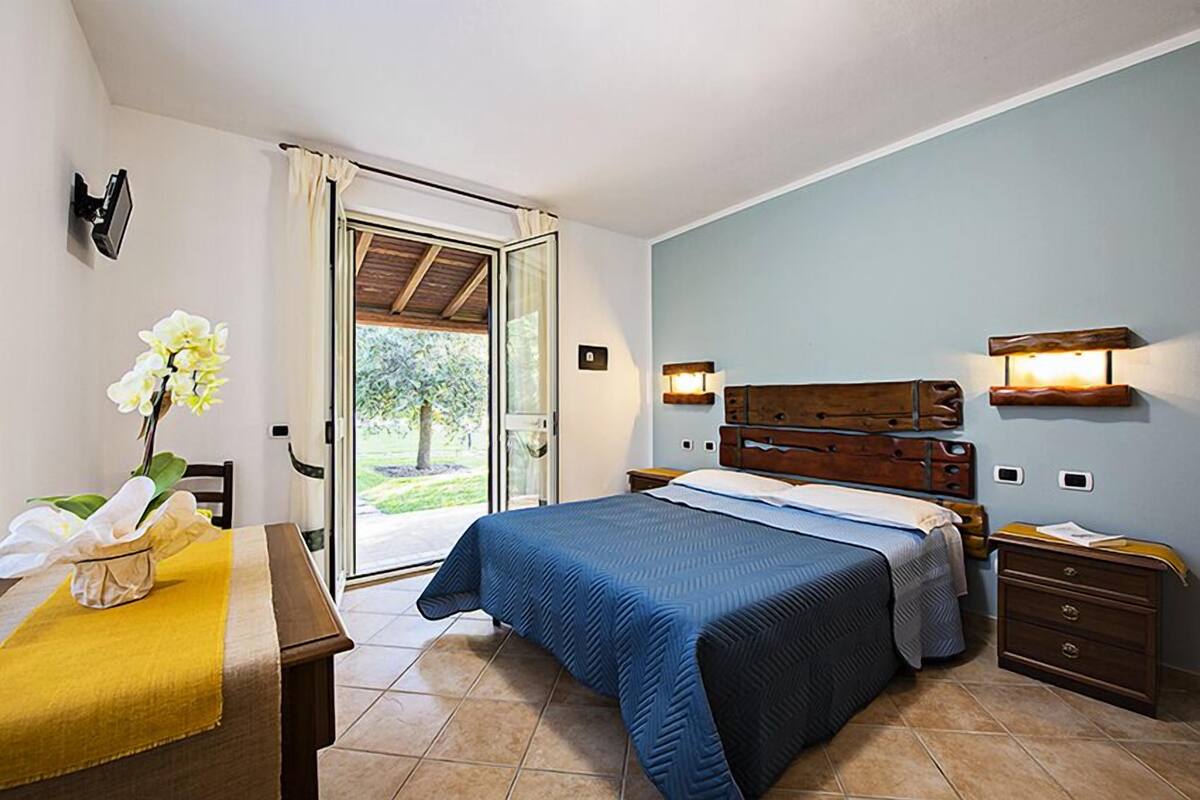 Ein Schlafzimmer mit Bett, Nachttischen, Schreibtisch mit Stuhl, Fenster mit Vorhängen und Blick ins Freie.