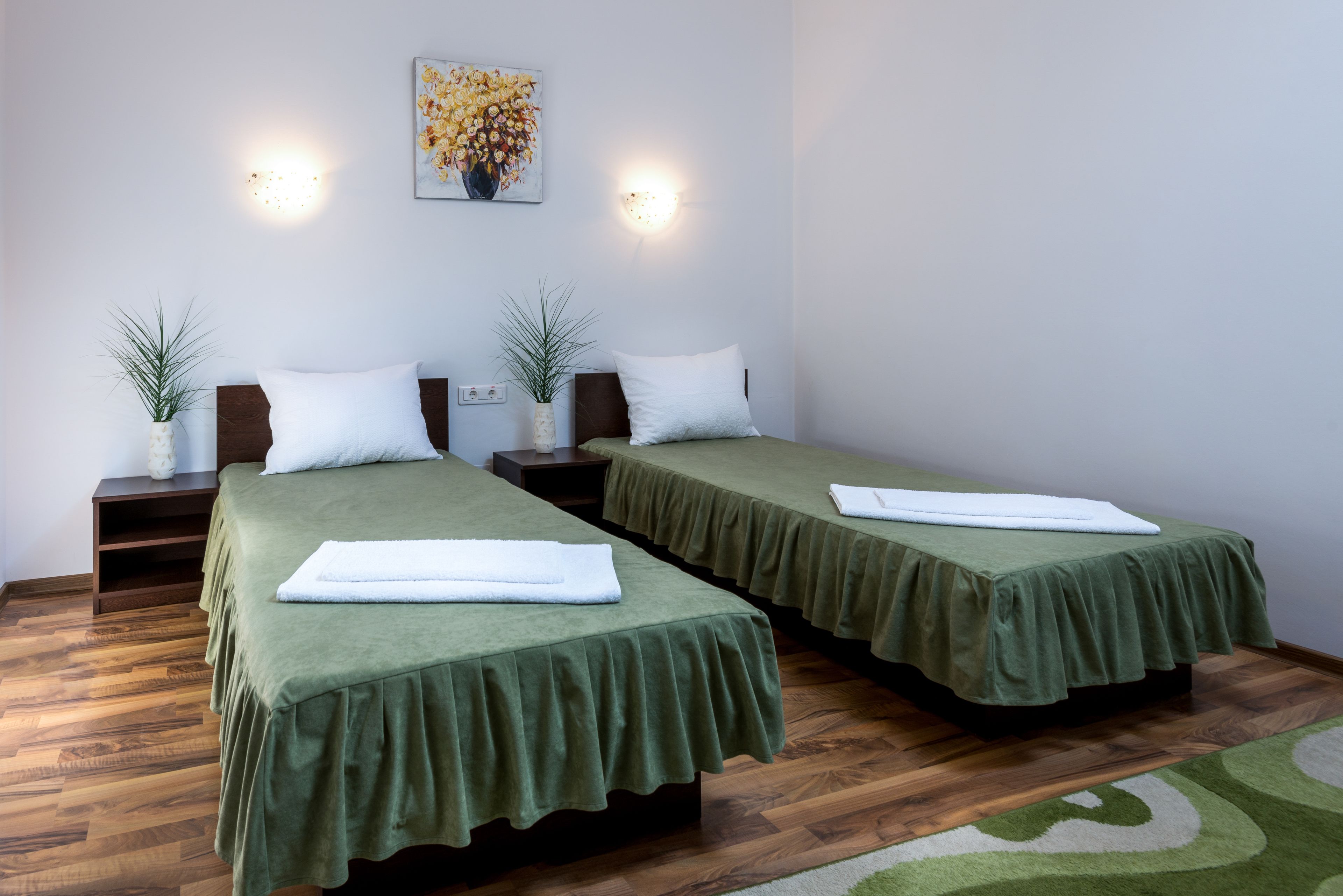 Hotellihuoneessa on kaksi erillistä sänkyä, joissa on vihreät päiväpeitteet ja valkoiset tyynyt. Huoneessa on myös kaksi yöpöytää, joissa on lamput, seinään kiinnitetty valaisin ja kukka-aiheinen taulu.