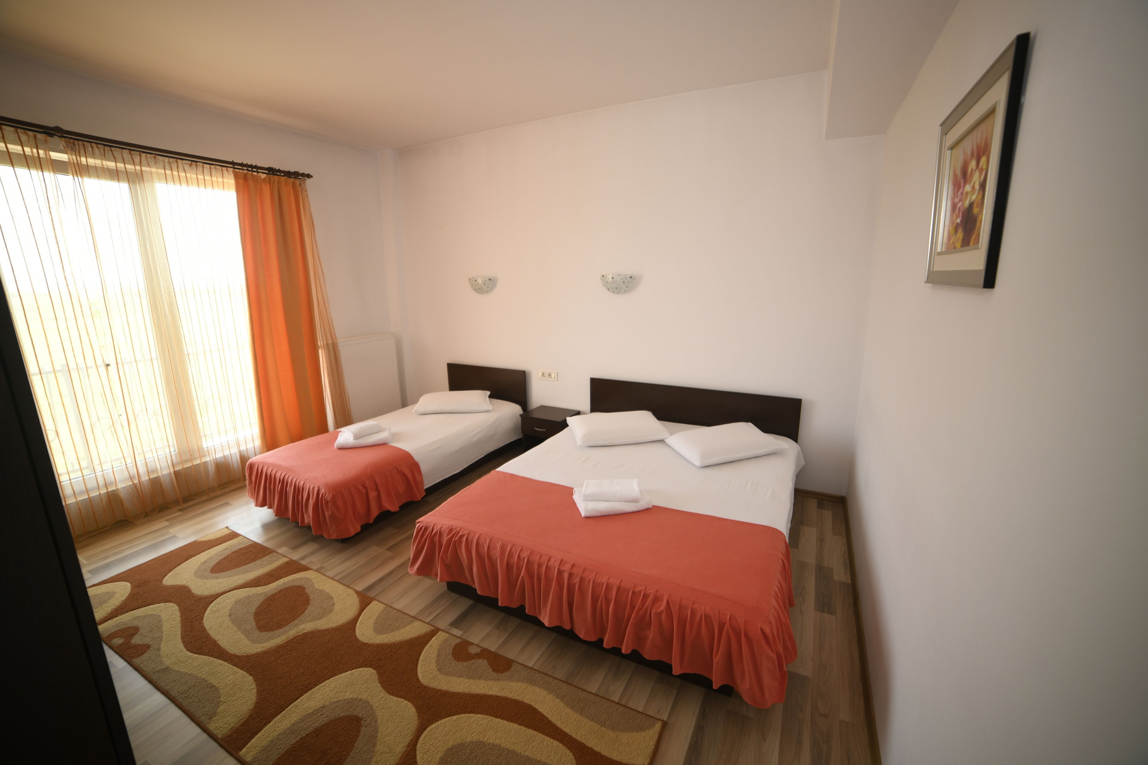A hotel room with two beds, a wooden floor, a patterned rug, and a window with curtains.
