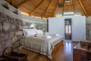 A bedroom with a stone wall, wooden ceiling, a bed with a quilt, and a suitcase on a wooden dresser. - The Windmill Adamas Sea View (Milos)