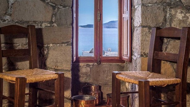 Una stanza rustica con due sedie di legno con sedili intrecciati, un muro in pietra e una finestra vista mare e montagne.