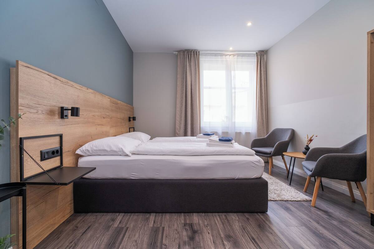 A modern hotel room with a wooden headboard, a large bed, a desk, two chairs, and a small table.