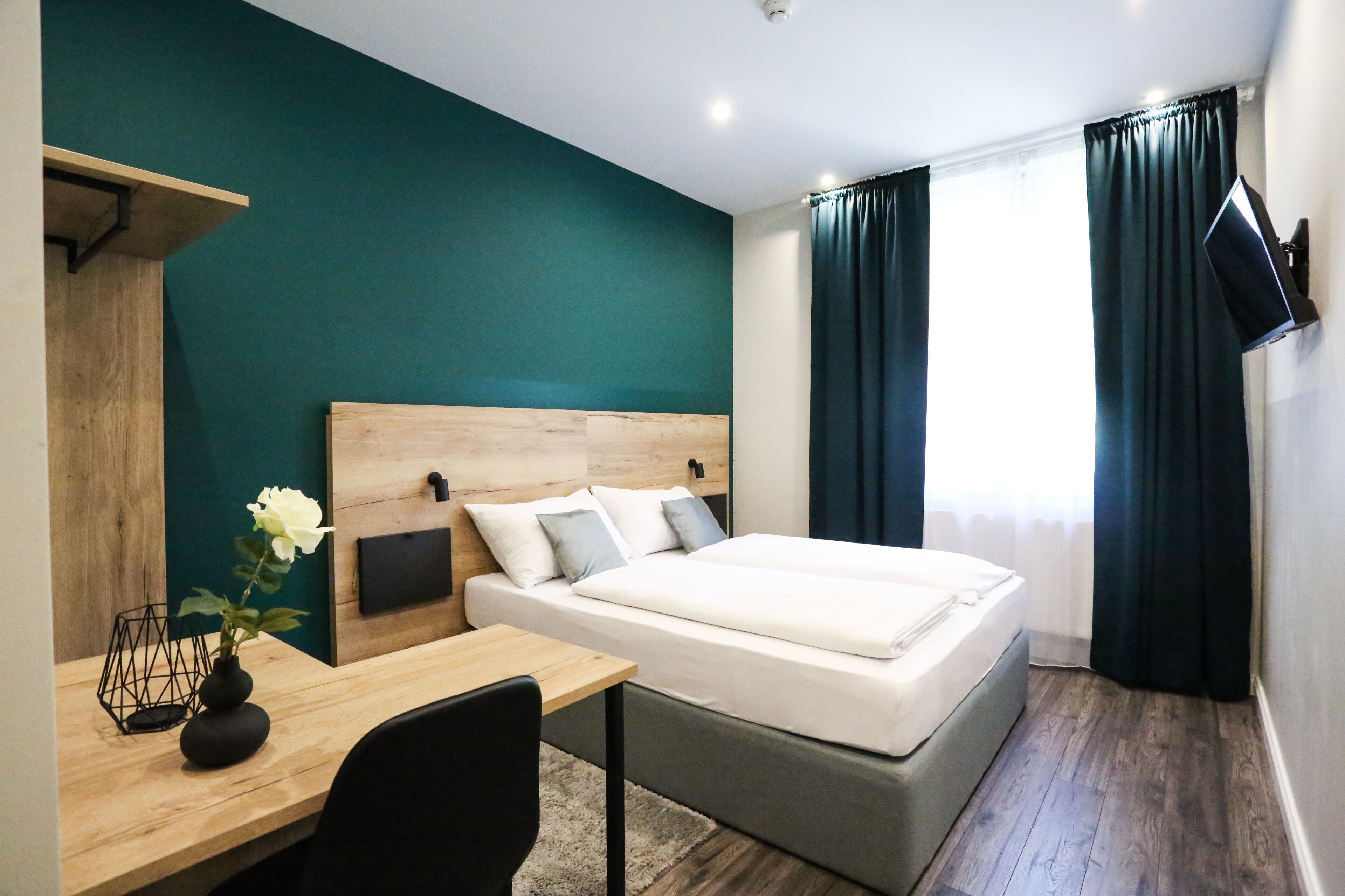 Una habitación de hotel moderna con cabecera de madera, un televisor de pantalla plana en la pared, un escritorio con un jarrón de flores y una cama grande con colchón gris.