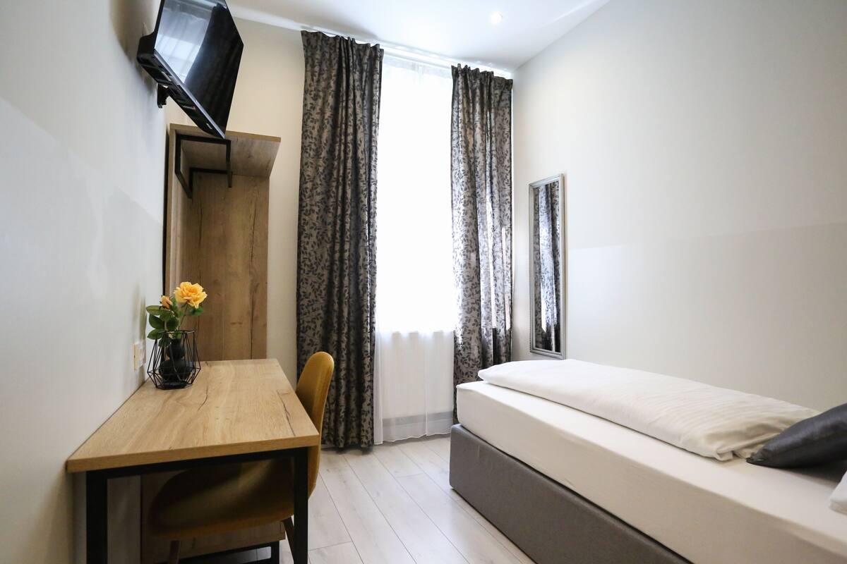 A compact room with a bed, a desk, a chair, a TV mounted on the wall, and a window with curtains.