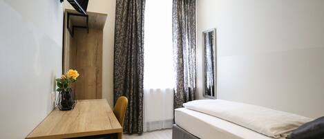 A compact room with a bed, a desk, a chair, a TV mounted on the wall, and a window with curtains.
