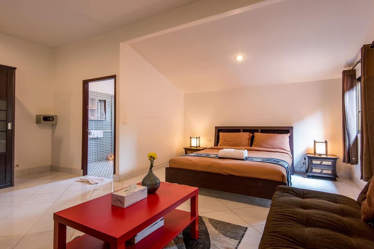 Habitación de hotel con cama, mesitas de noche, una mesa roja, un sofá marrón y vista a un edificio.
