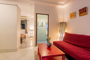 A modern living room with a red sofa, a wooden bench, and a red table with a vase. A door leads to a bathroom with a shower and a toilet visible. - Tananza Resort and Homestay Phuket (Rawai)