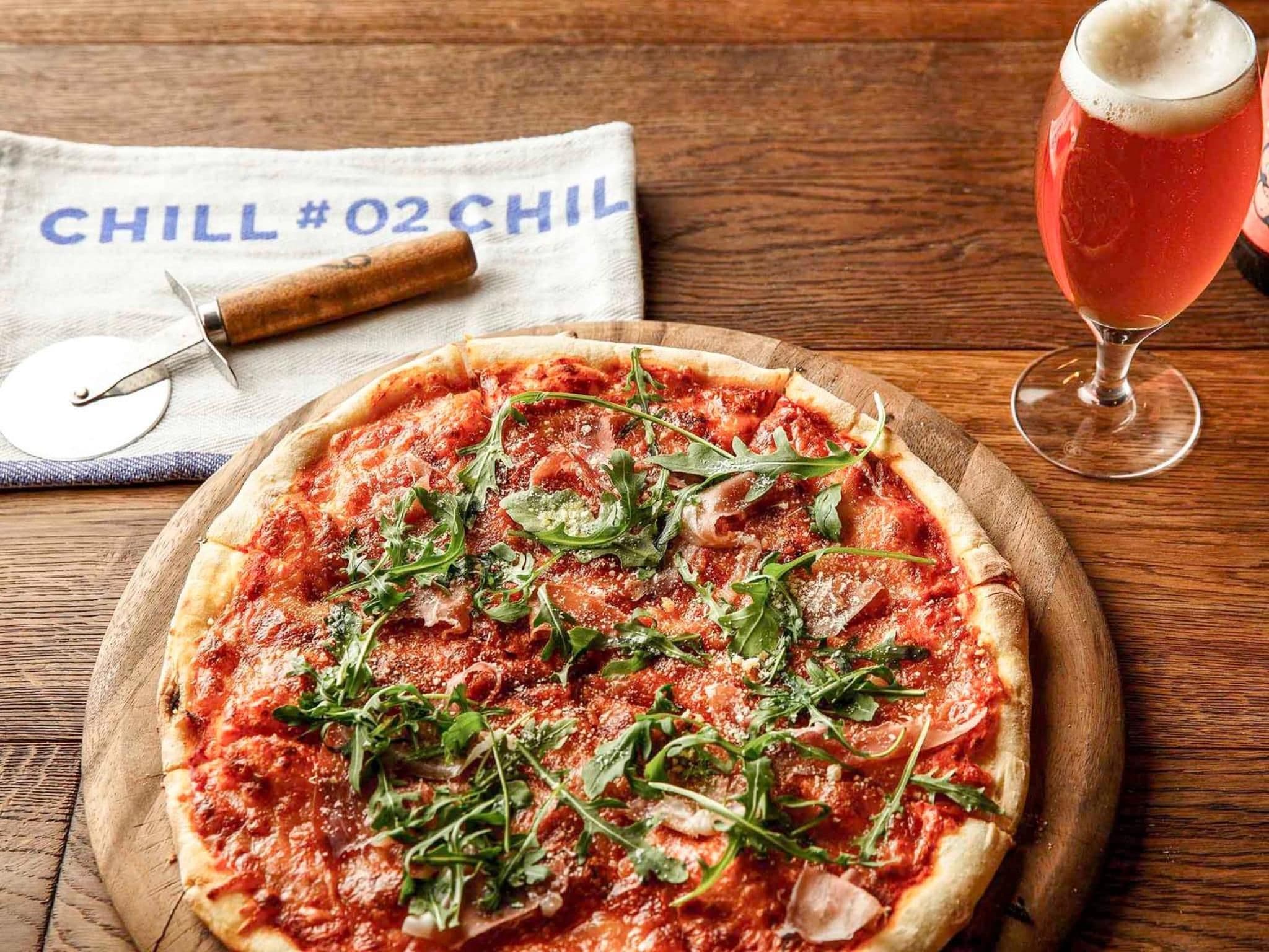 Una pizza con verduras y queso sobre una tabla de madera, un cortador de pizza y una bebida en un vaso sobre una mesa de madera.