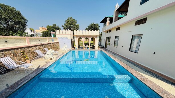 A rectangular swimming pool with clear blue water, surrounded by a stone wall and a white building with a balcony. - Sajjan Bagh Resort (Pushkar)