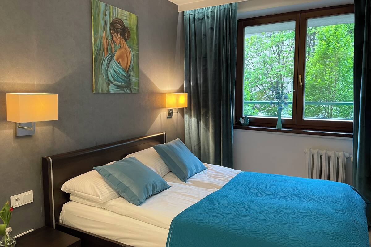 Una camera d'albergo con un letto, due lampade a parete, un quadro, una finestra con tende e un termosifone.