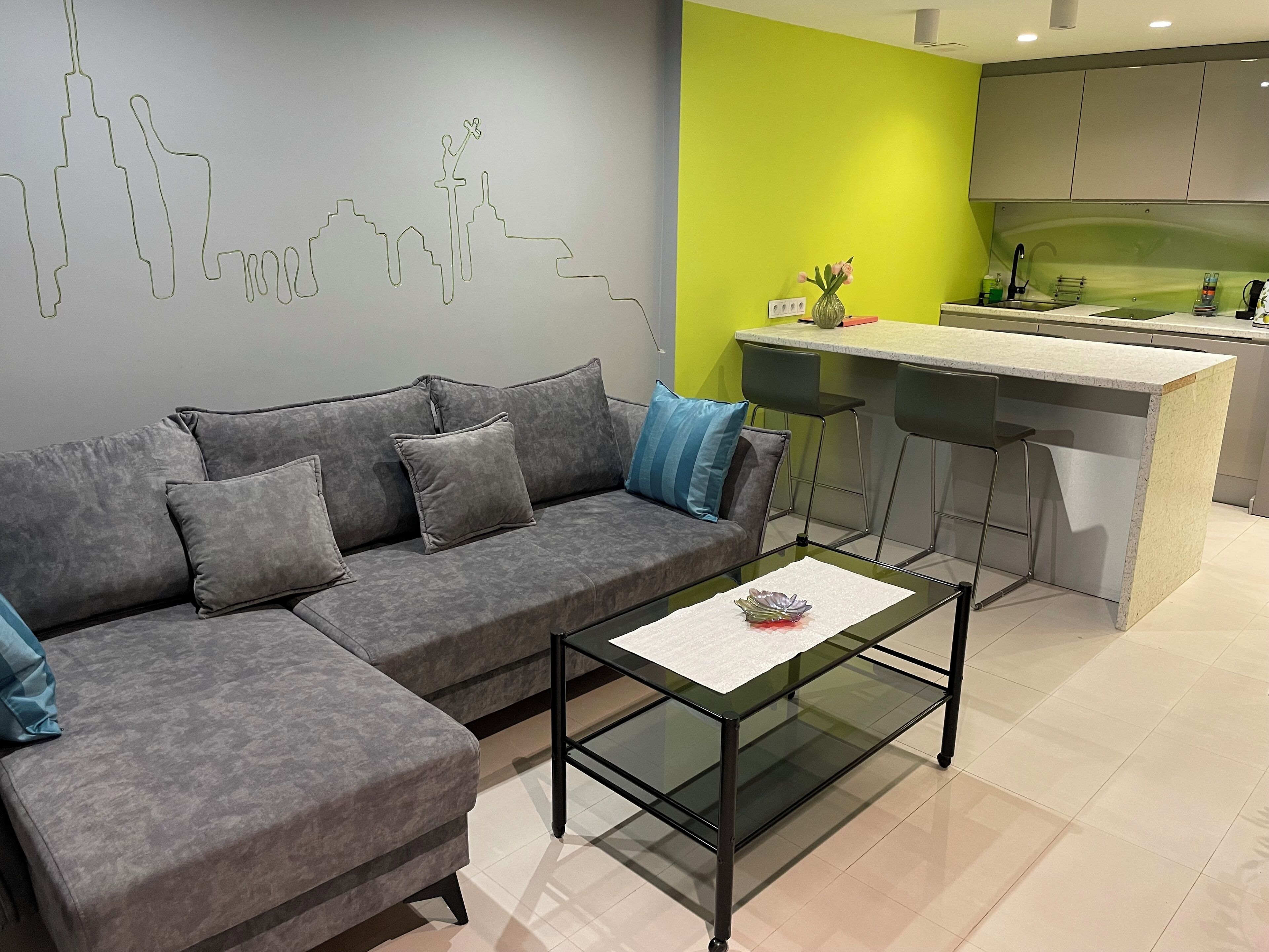 Ein modernes Wohnzimmer mit einer grauen Couch, einem Glastisch und einer Küchenzeile mit einer grünen Akzentwand.