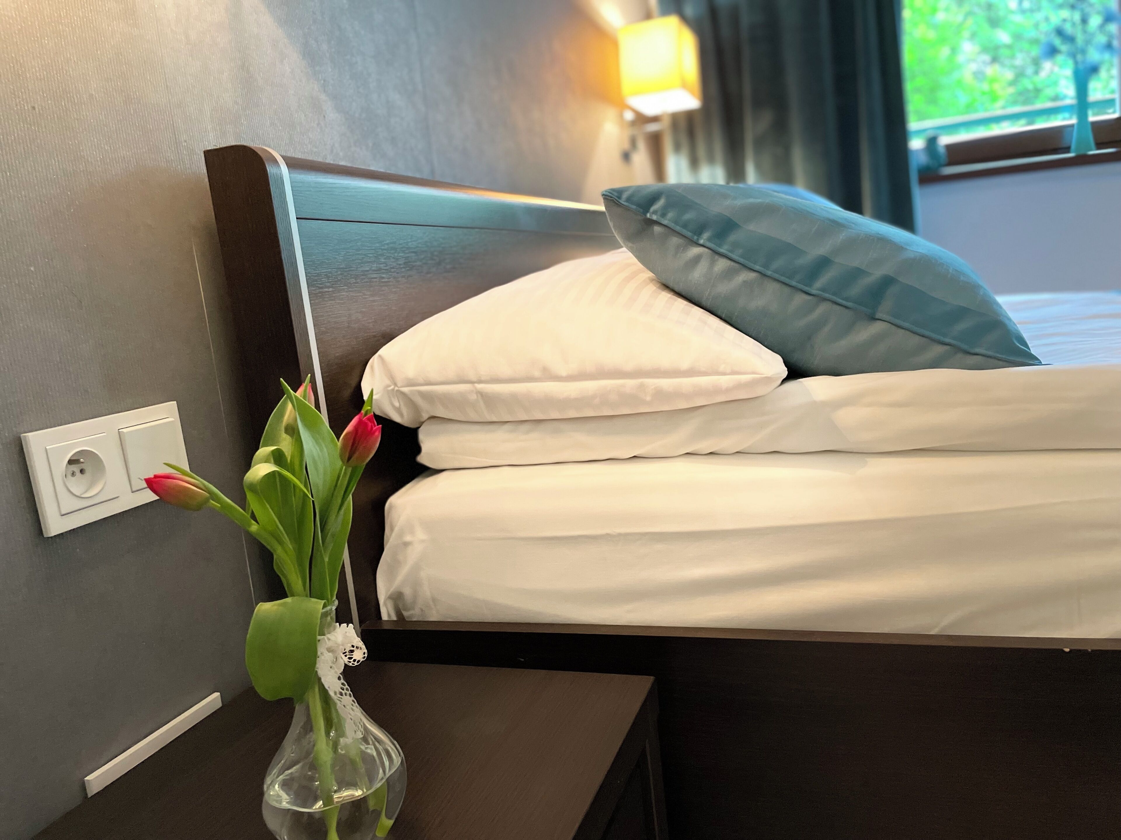 A neatly made bed with white and blue pillows, a clear vase with a flower, and a wall socket.