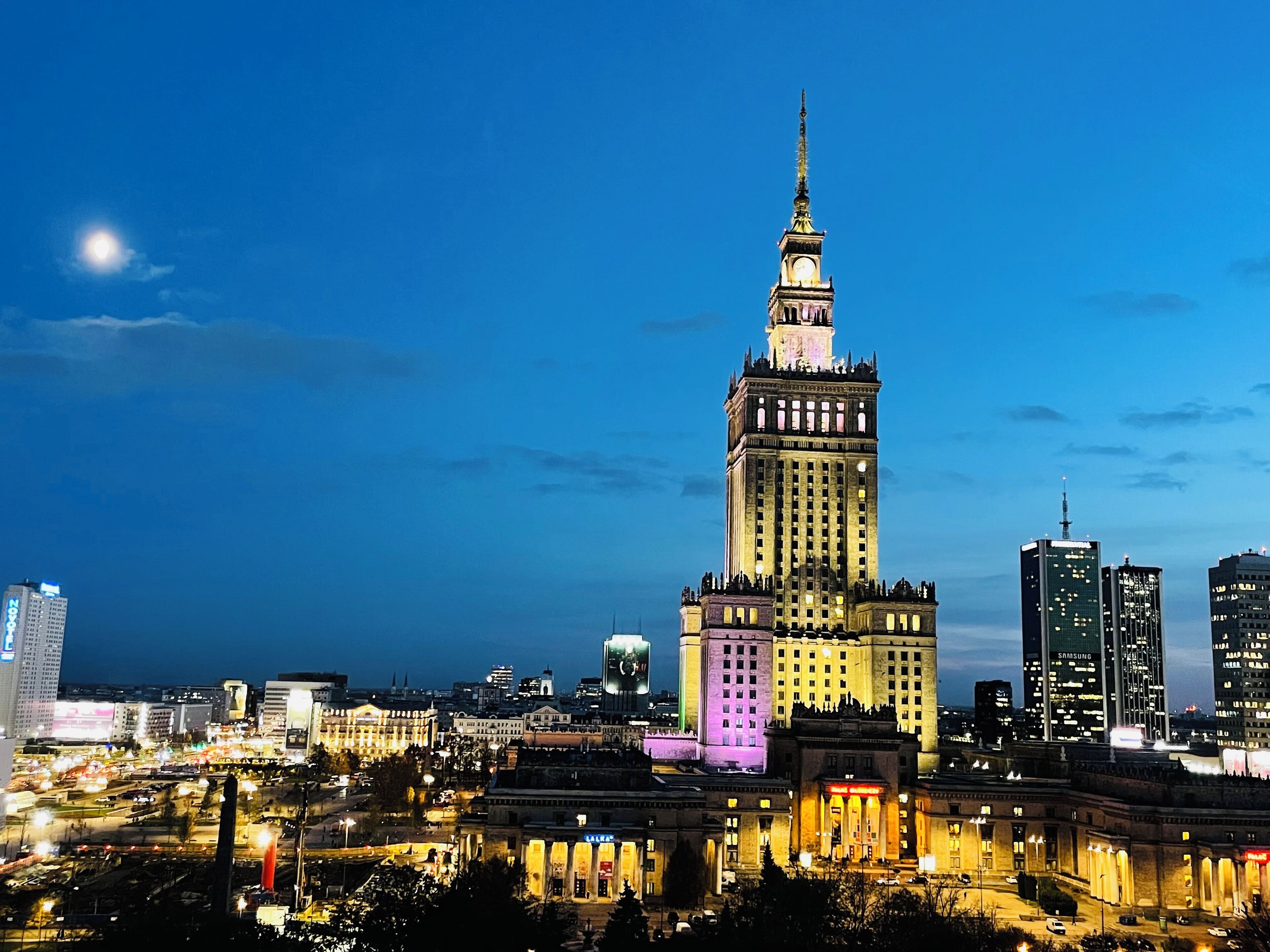 Un edificio histórico con una torre de reloj, rodeado de modernos rascacielos e iluminado por farolas.