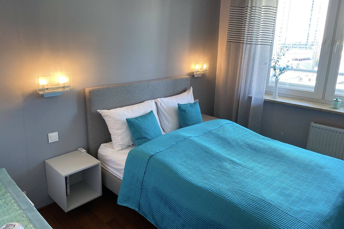 Un letto rifatto con una coperta e dei cuscini azzurri, un comodino con una lampada e una finestra con tende.