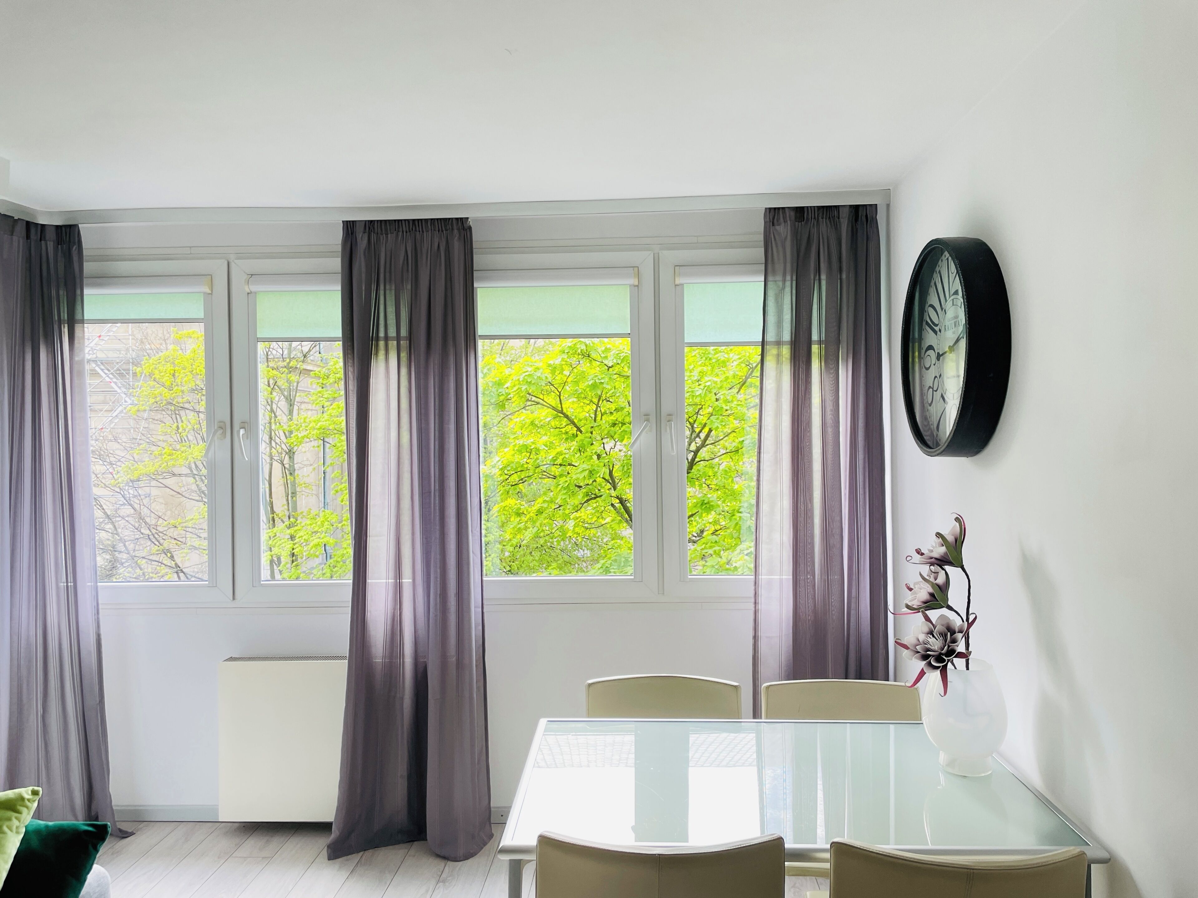 A room with a dining table, chairs, a clock, and a window with curtains.