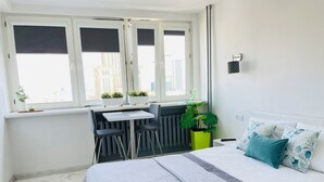 A bedroom with a bed, a desk with two chairs, a radiator, a window with blinds, and a potted plant. - Good Time Apartments (Warsaw)