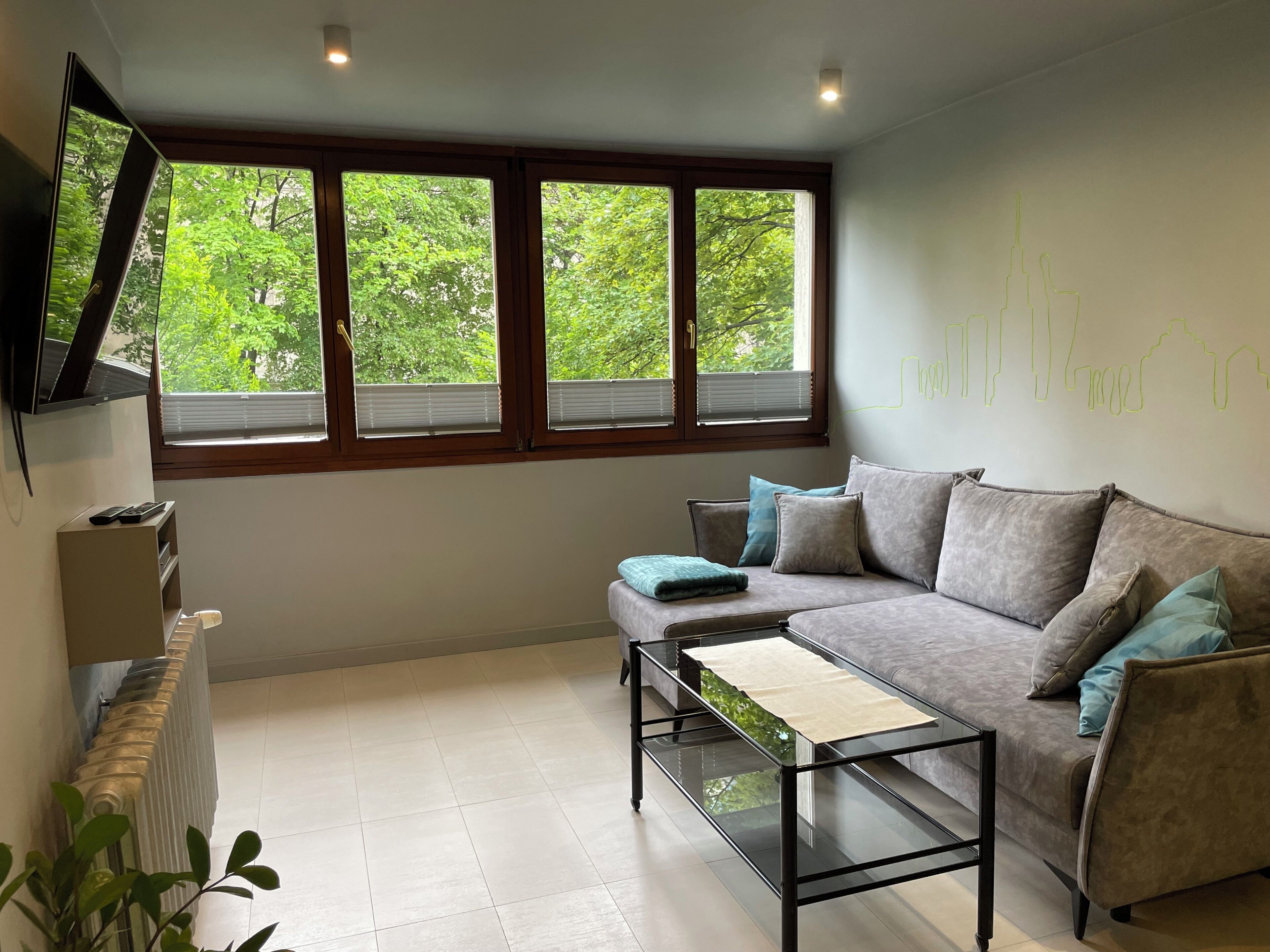 A modern living room with a sofa, a glass coffee table, a TV mounted on the wall, and a window with blinds.