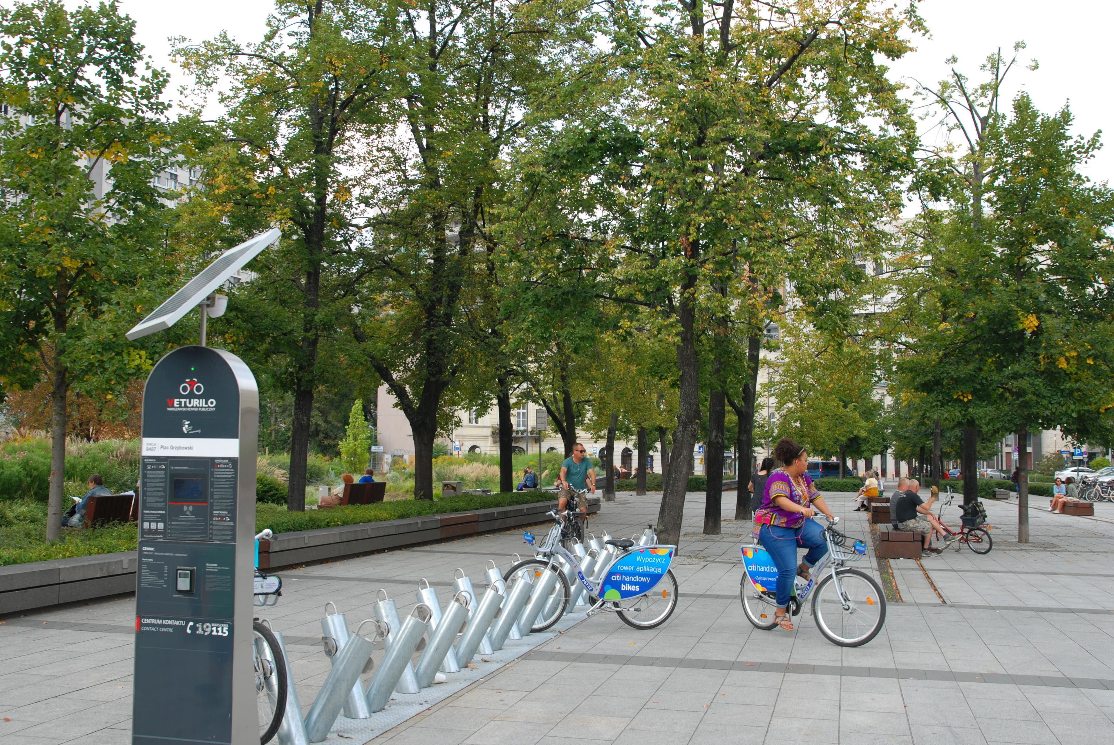 Een openbaar fietsdeelsysteem station met een zonnepaneel, omgeven door bomen en mensen.
