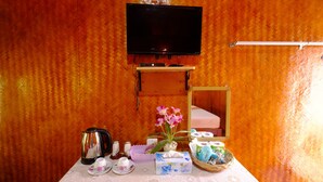 Una habitación con un televisor montado en la pared, un pequeño estante de madera con un altavoz, una mesa con juego de té, una cesta de artículos de aseo y un espejo.