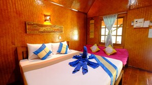 Habitación de hotel con paredes de madera, cama con ropa de cama azul y blanca y una colcha rosa, y una ventana con cortina.