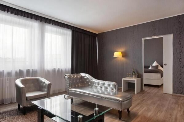 Una sala moderna con un sofá, un sillón y una mesa de centro de vidrio. Hay una lámpara en la pared y una cama al fondo.