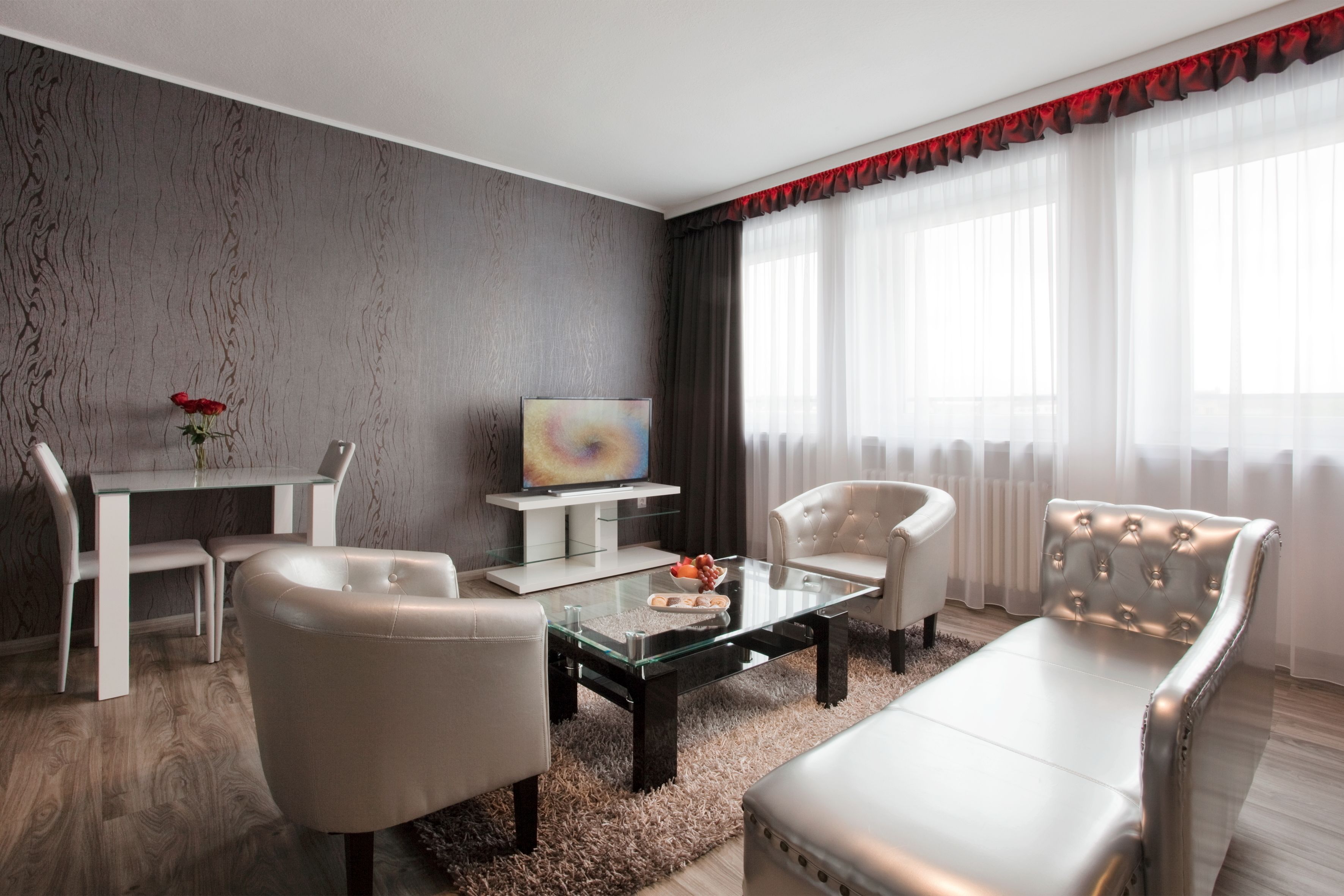 Een moderne woonkamer met een glazen salontafel, twee witte fauteuils, een bank, een eettafel met stoelen, een flatscreen televisie en een decoratieve vaas met bloemen.