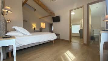 Quarto com teto de madeira, cama com roupa de cama branca, escrivaninha com luminária e televisão fixada na parede.