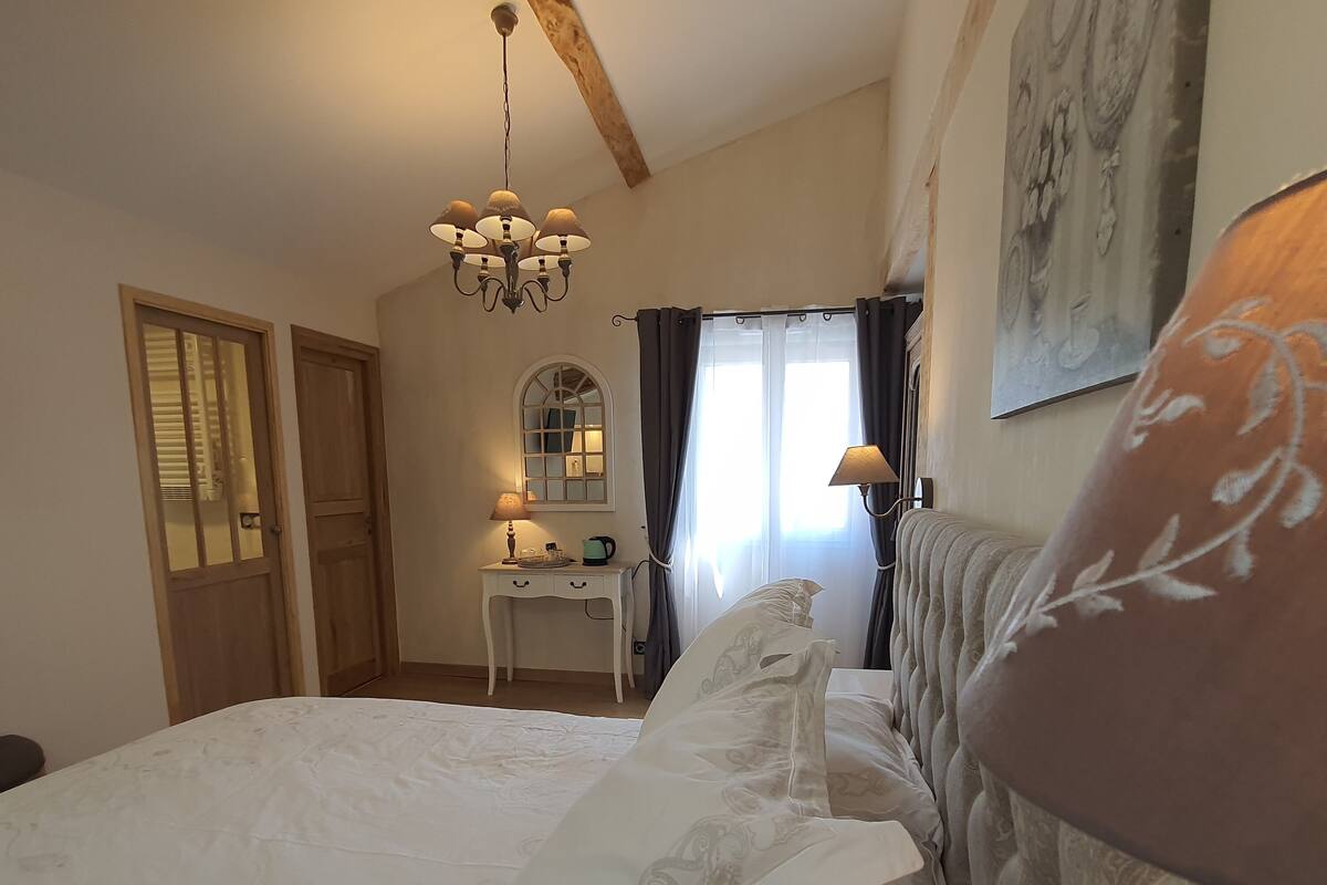 Ein Schlafzimmer mit einem großen Bett, einem Schreibtisch inklusive Spiegel, einer Chandelüster und einem Fenster mit Vorhängen.