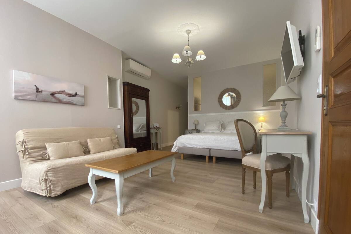 Ein Schlafzimmer mit einem Bett, einem Nachttisch aus Holz, einem Schreibtisch mit Lampe, einem Sessel, einer Couch, einem Holzboden, einer Deckenleuchte und einem an der Wand befestigten Fernseher.