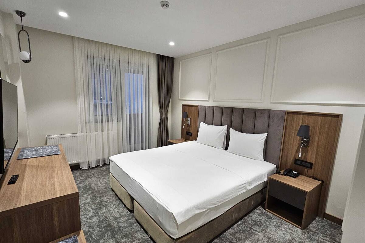 Una habitación de hotel moderna con una cama grande, un escritorio de madera, una mesita de noche y un ventanal con cortinas translúcidas.