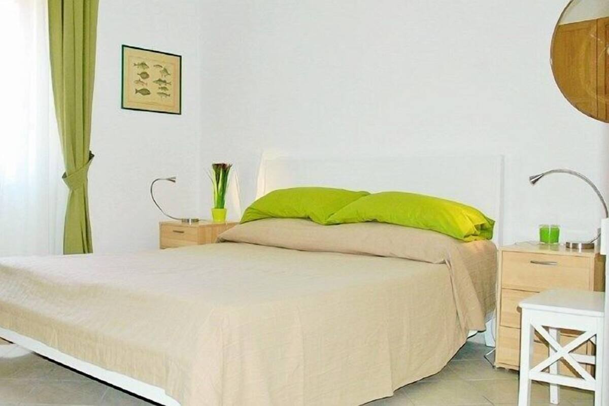 A bedroom with a bed, a bedside table, a nightstand, a lamp, a picture on the wall, and a window with curtains.