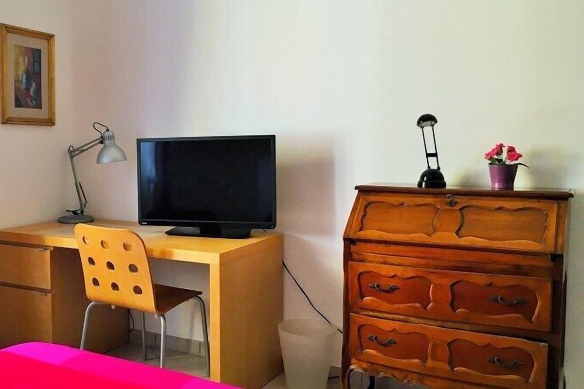 A room with a wooden desk, a computer monitor, a chair, a dresser, and a window with red curtains.