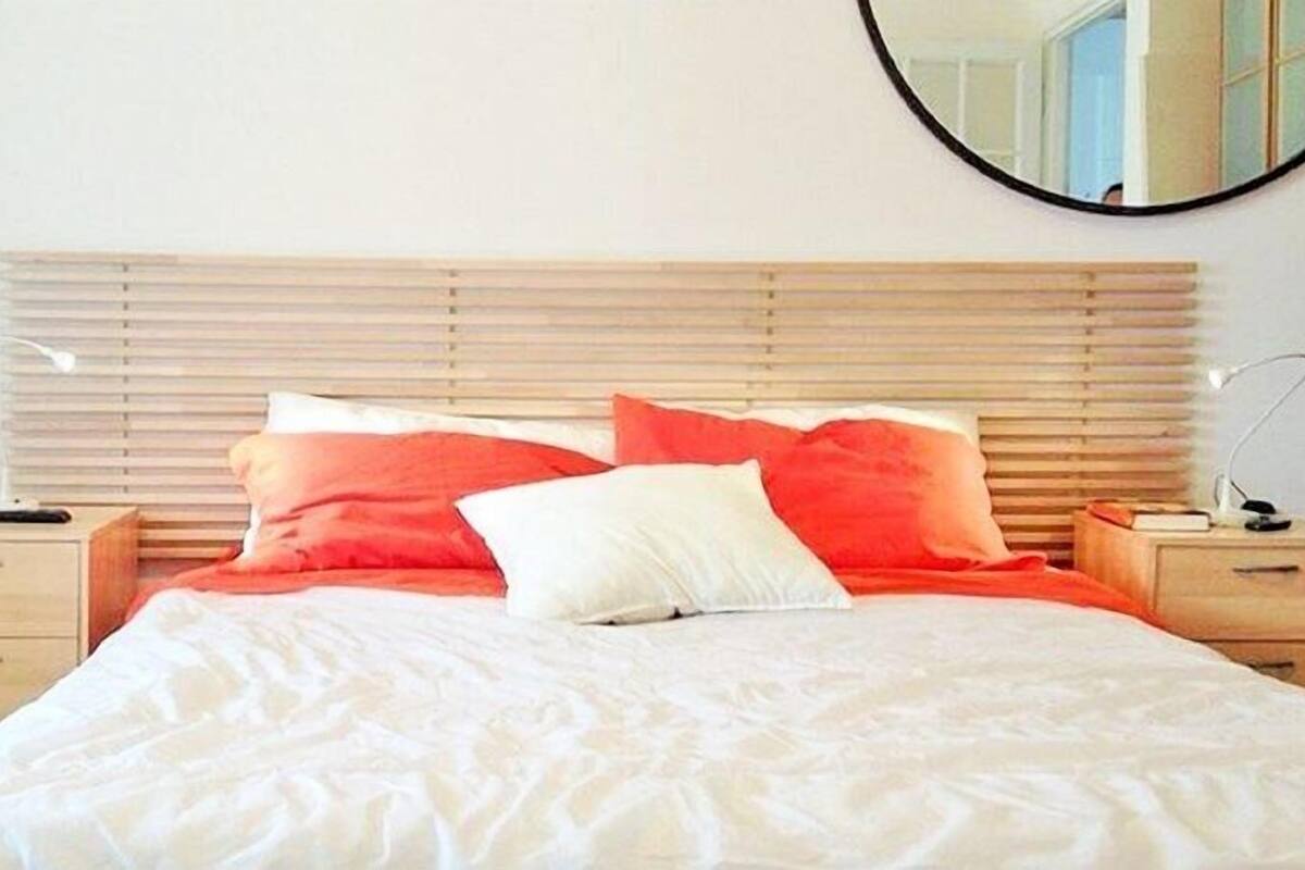 A neatly made bed with white and red bedding, wooden headboard, bedside tables, and a round mirror on the wall.