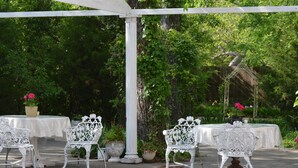 An outdoor patio with white metal chairs and tables, surrounded by lush greenery and a pergola structure. - The Carleton House (Bonham)