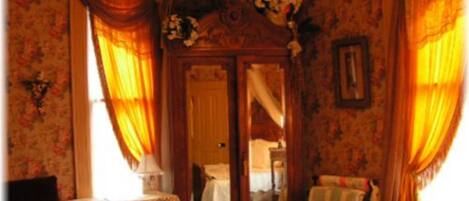 Una habitación con papel tapiz floral, un armario de madera, una mesa con mantel de encaje y cortinas.
