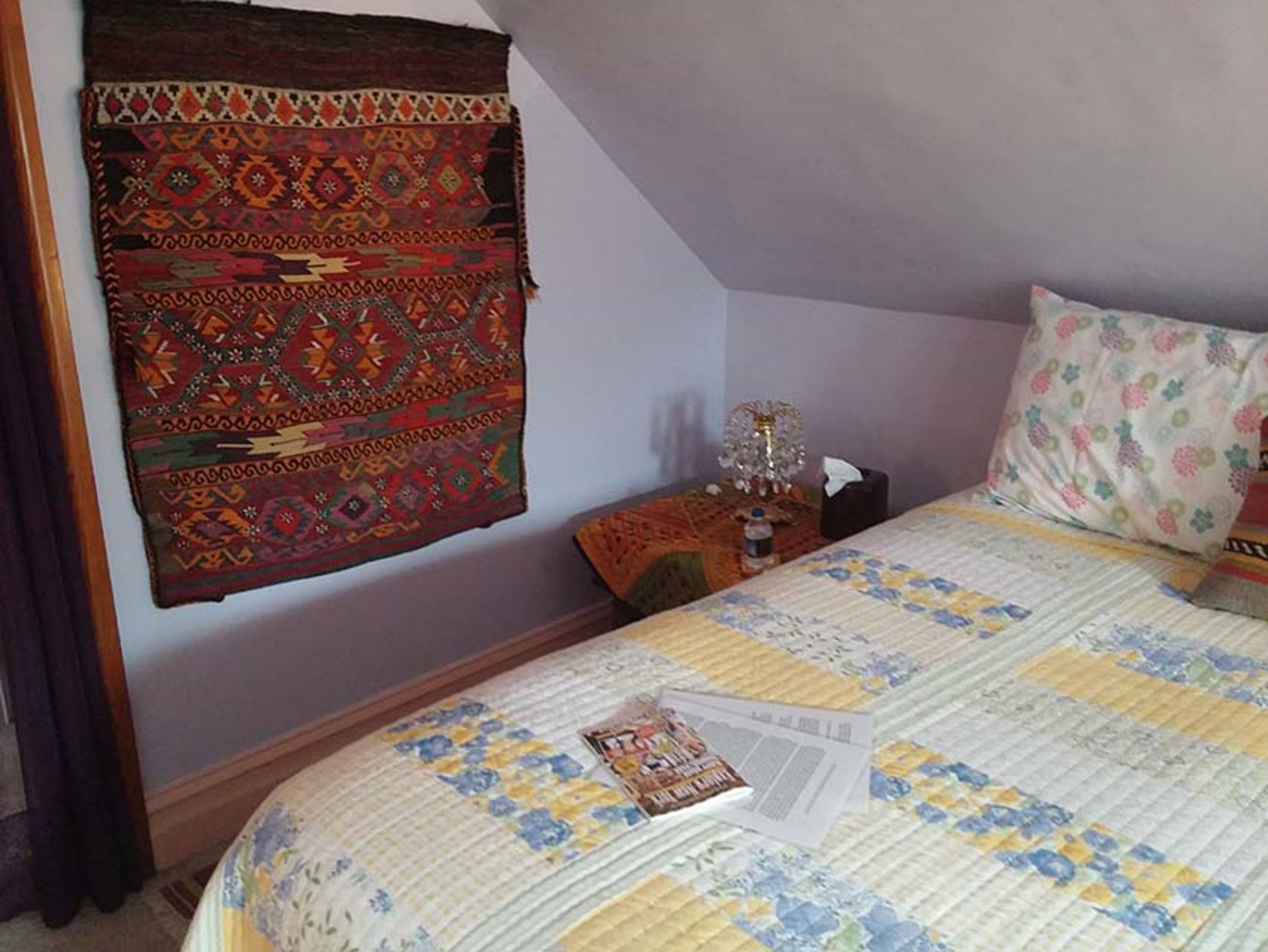 A bedroom with a bed, a patterned wall hanging, a nightstand with a lamp, and a floral pillow.