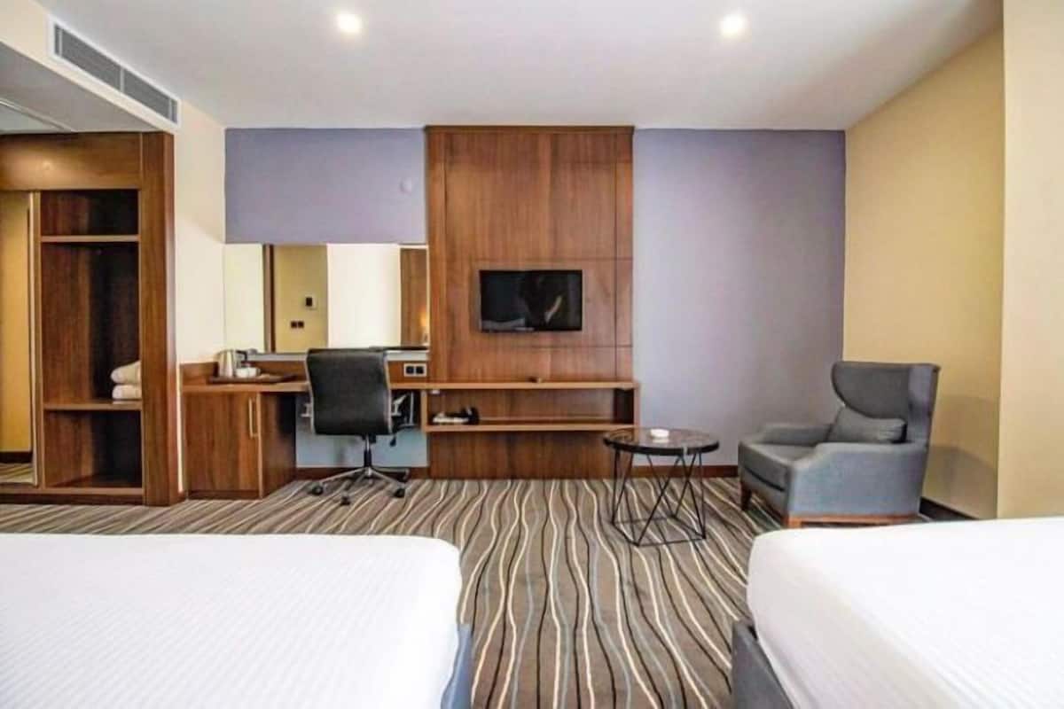 A hotel room with two beds, a desk with a chair, a small table, a TV mounted on the wall, and a wardrobe.
