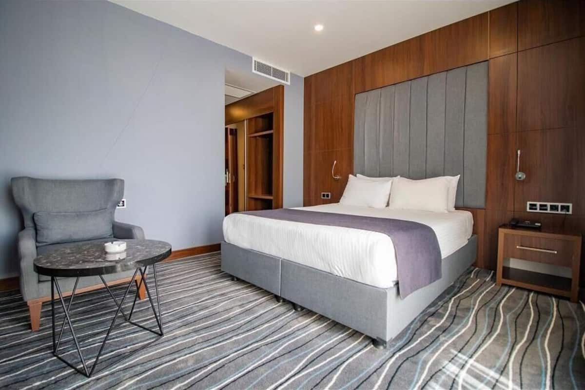 A hotel room with a bed, a grey armchair, a small round table, and a wooden wardrobe.