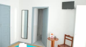 A room with a door, a mirror, a small table with a clock, a chair, and a TV mounted on the wall. - Peneco Albufeira GuestHouse - Hostel (Albufeira)