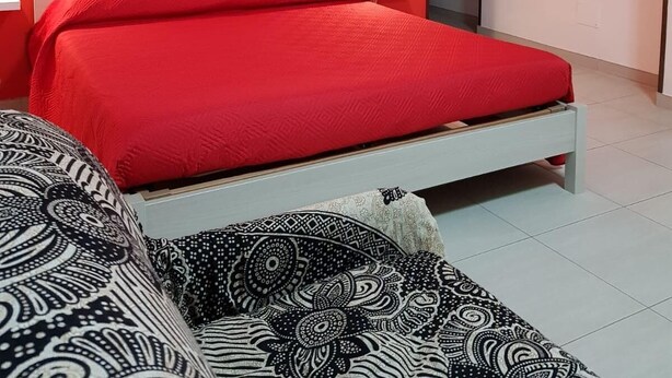 A modern bedroom with a red wall, a white bed, patterned pillows, a clock, and wall-mounted lights. - Monolocale Rosso (Turin)